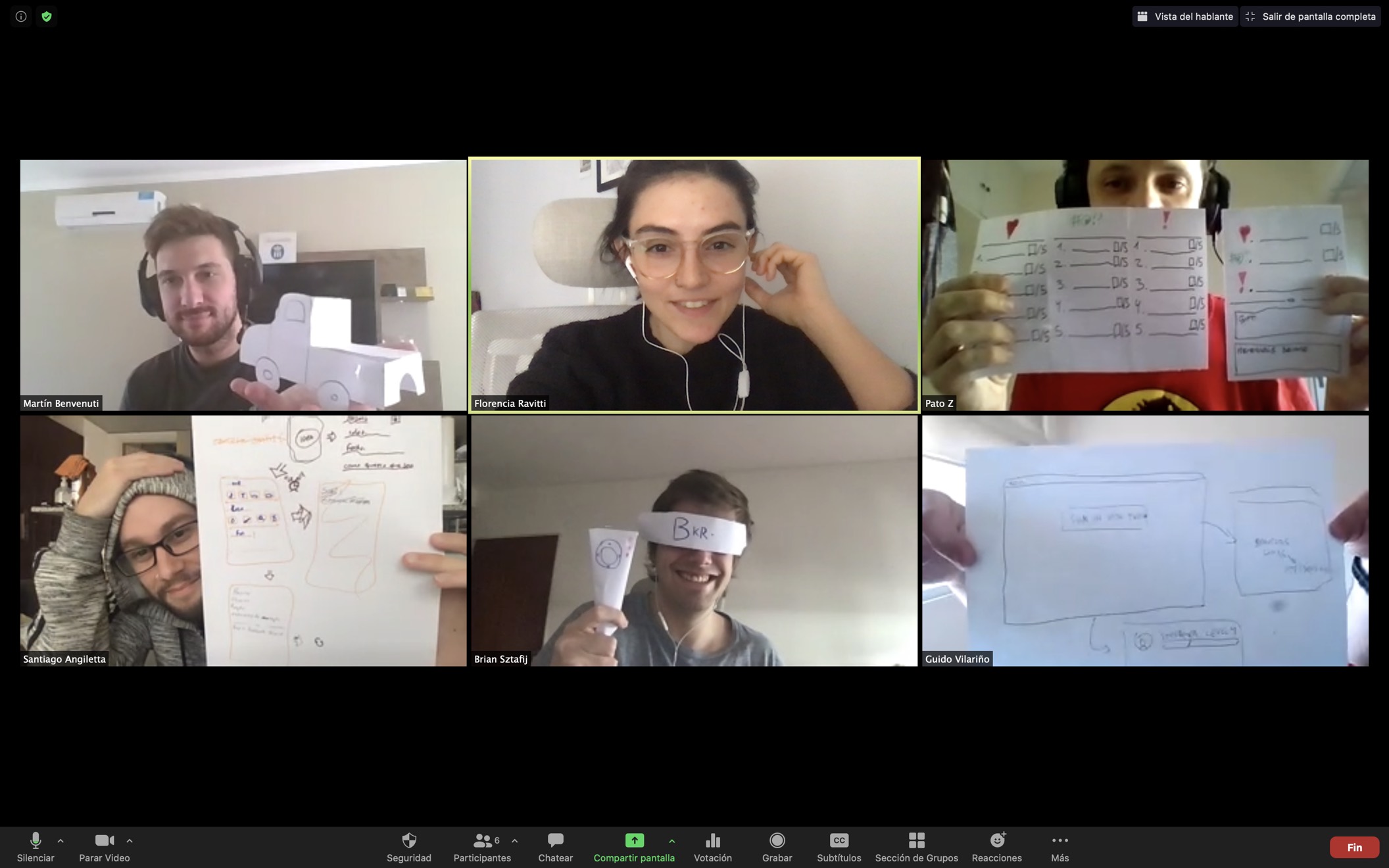 A Zoom grid of six participants in a session, each holding a paper
              prototype up to the camera: a cardboard truck, hand-drawn app
              screens, a checklist and a paper headband.