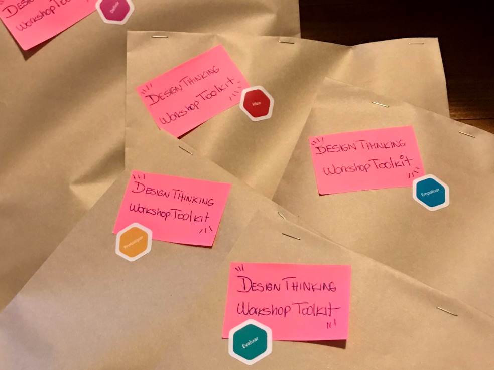 Kits packed in brown paper envelopes, each stapled shut and labelled
              with a pink hand-lettered card reading Design Thinking Workshop
              Toolkit, plus a coloured sticker for one of the five stages.